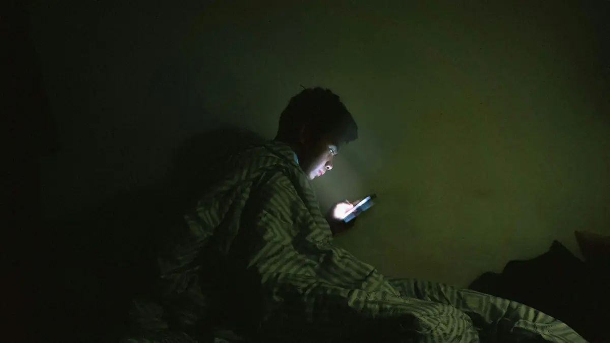 Homem enrolado no cobertor olhando o celular no escuro.