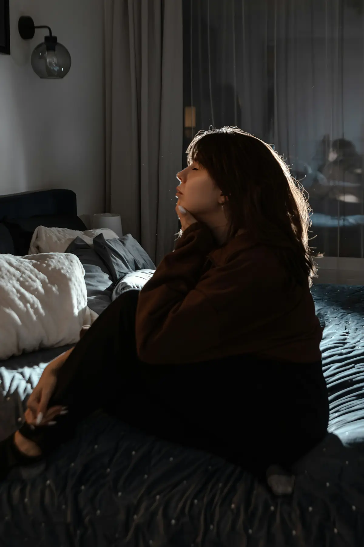 Jovem sentada na cama, pensativa, em um quarto de luz baixa.